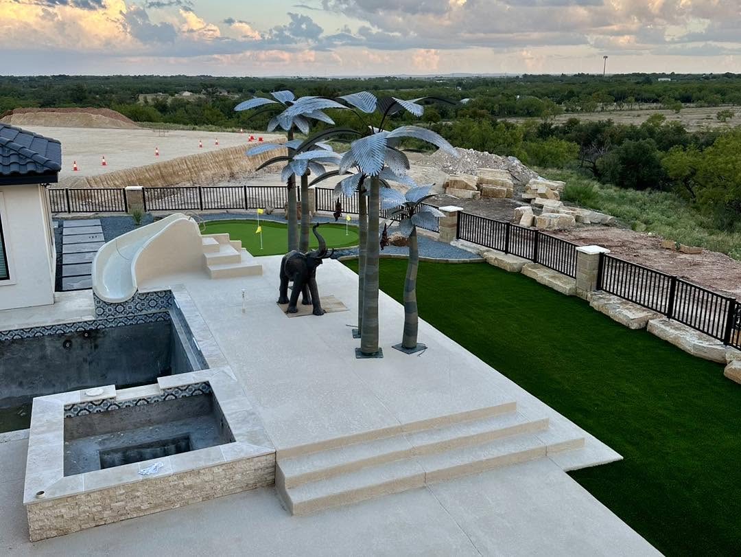Aerial view of a hilltop backyard with pool, water slide, putting green, turf, and metal palm trees