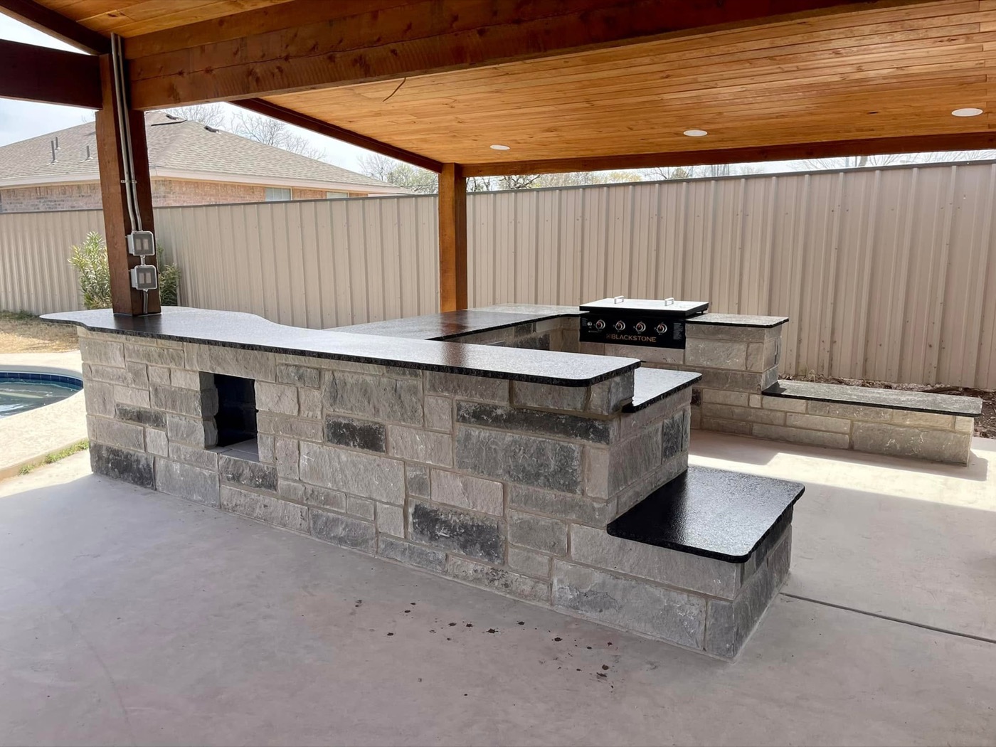 Poolside outdoor kitchen with gray stone base, black granite counters, and griddle under a wood-ceiling pavilion