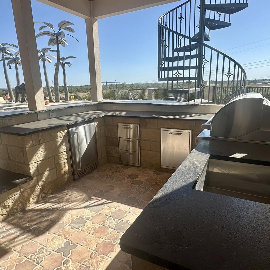 Outdoor kitchen with limestone cabinetry, stainless appliances, and patterned tile floor beside a spiral staircase