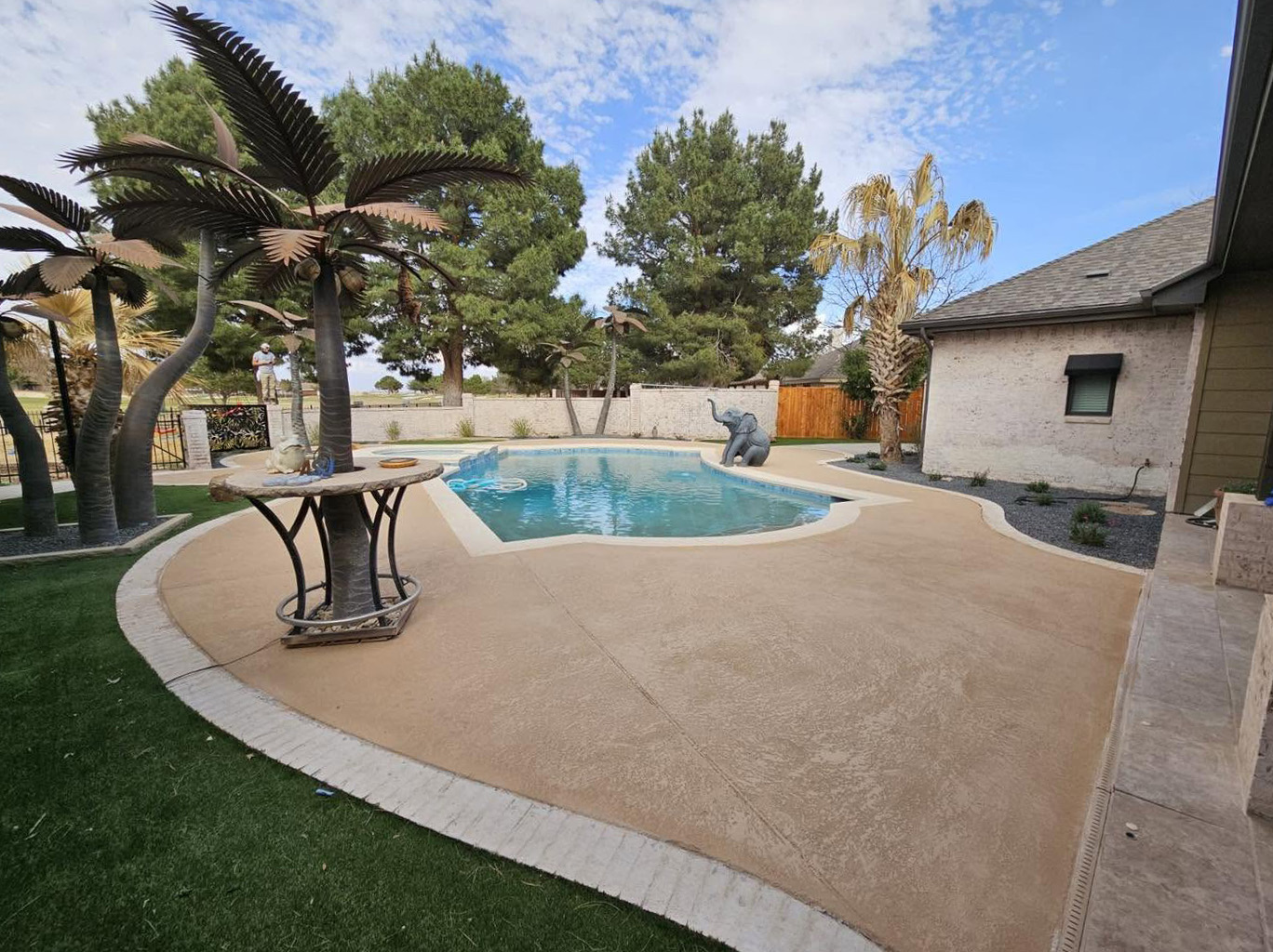 Renovated pool deck in warm tan finish with sculpted metal palm trees and turf border
